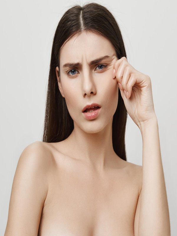 Wrinkles attacking her beautiful face. Studio shot of upset irritated young european woman, standing naked while pulling face near eye and showing aging results, being sad about it