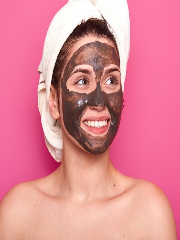 Indoor shot of young attractive woman with white towel on her head, has naked body, smilling isolated over pink background in studio, looks aside, having chocolate mask on her face. Skincare concept.
