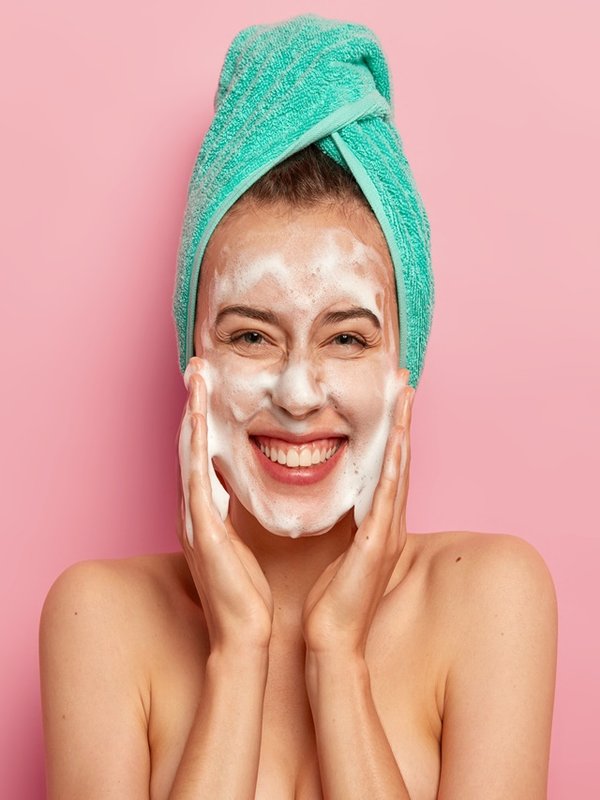 Pampering and hygiene concept. Happy young European woman massages cheeks, aplies bubble foam, washes face, smiles positively, has naked body, enjoys taking shower, wants to have clean skin.