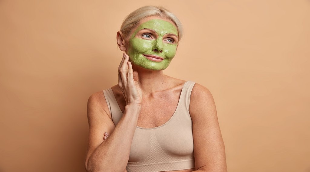 Portrait of dreamy middle aged woman applies green mask on face stands thoughtfully and looks away undergoes beauty procedures dressed casually isolated over beige background. Skin care concept