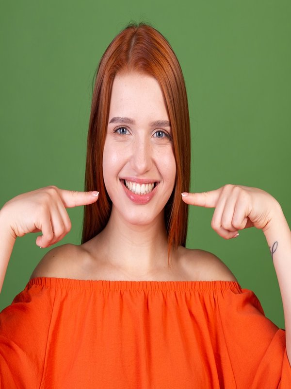 Young red hair woman in casual orange blouse on green background point fingers on white teeth perfect smile