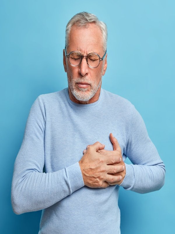 Senior man in spectacles presses hand to chest has heart attack suffers from unbearable pain closes eyes wears optical glasses poses against blue background. People age and problems with health