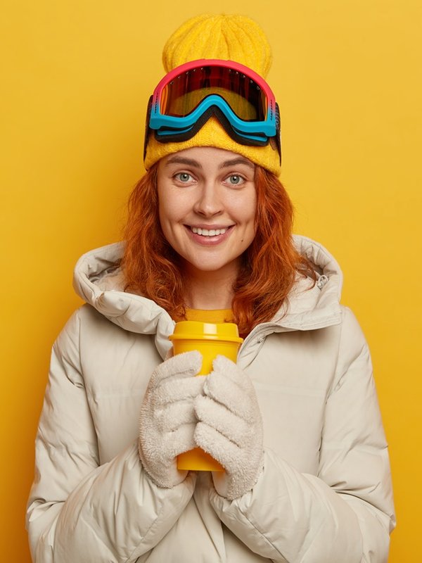 Woman skier wears warm winter outerwear, holds yellow takeaway cup with hot tea, wears cap and ski goggles, smiles pleasantly, models indoor. Skiing, sport, active lifestyle and people concept