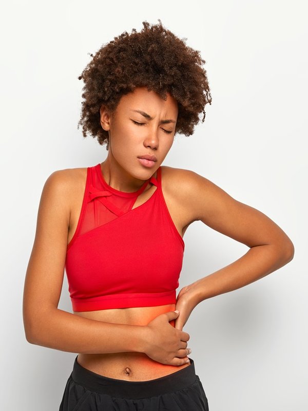 Dissatisfied African American woman holds aching hip, has kidney inflammation, touches location of pain near ribs marked with red dot, wears sport bra, shows perfect figure, has sporty shaped body