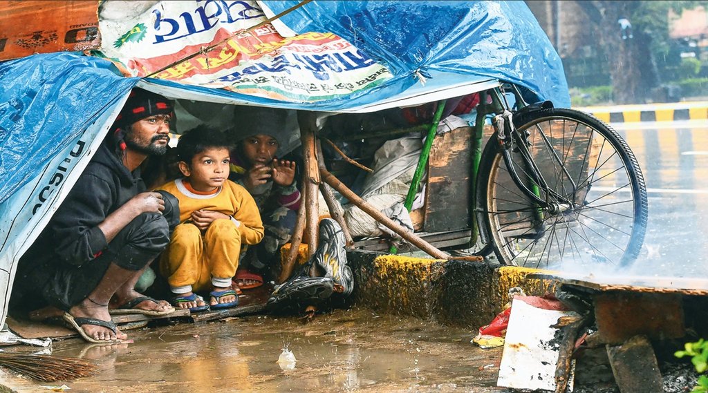 राजधानी दिल्ली में शनिवार को बारिश के दौरान सड़क किनारे अपने अस्थायी टेंट में बैठा एक परिवार। 