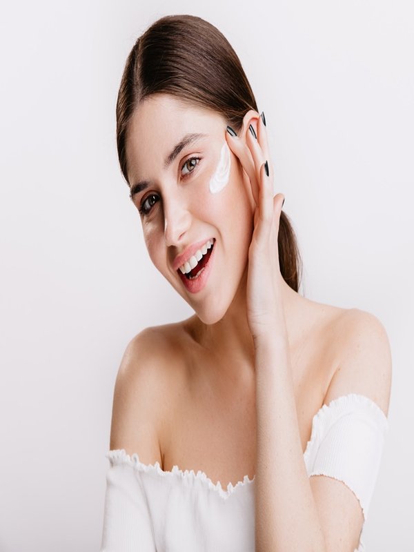 Smiling green-eyed girl puts cream on clean face. Brunette in white top posing on isolated background.