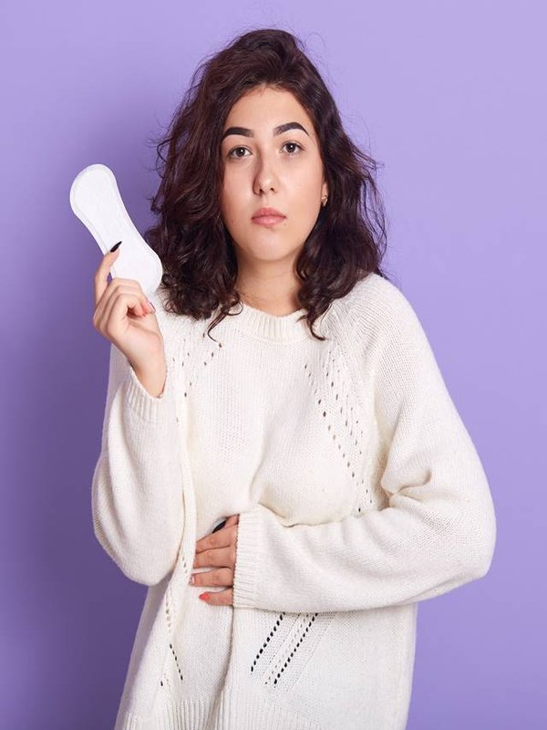 Gloomy Caucasian woman cares about personal hygiene, holds sanitary pad in hands, suffers from pain during menstruation, has sad expression, wear white outfit, poses in studio isolated over lilac wall