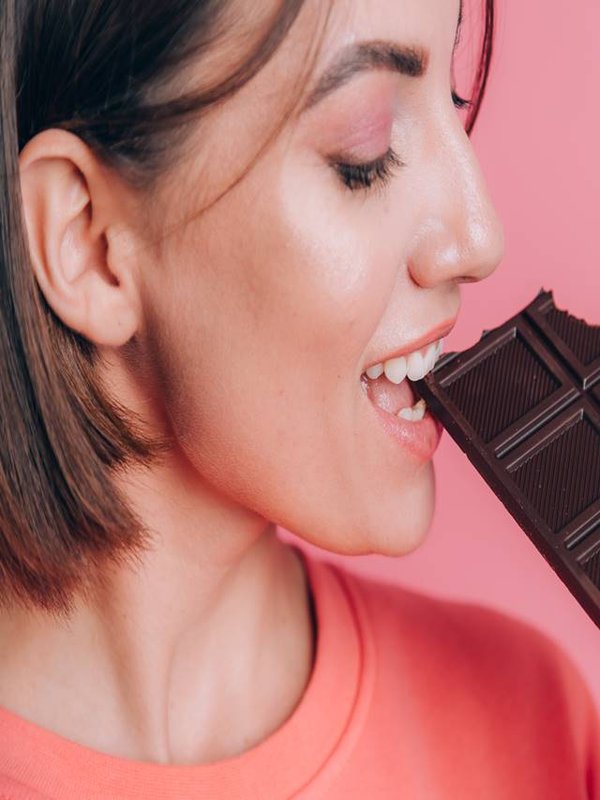Beautiful young happy woman with a bar of chocolate on a pink background and bright makeup, a close-up frame takes a bite
