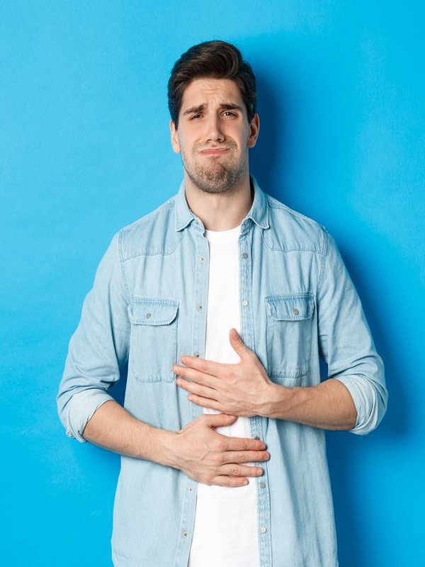 Man holding hands on belly and grimacing from pain, complaining on stomach ache, standing against blue background