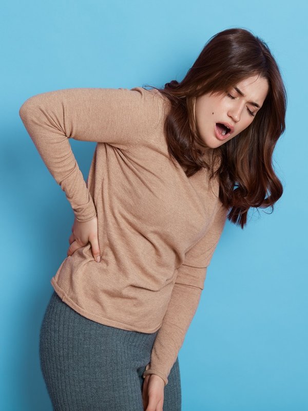 Dark haired European female wearing casual attire, having strong pain in back, keeping hands in her body, girl with widely opened mouth, suffering from illness.