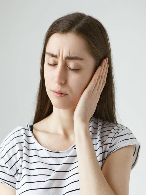 Studio shot of attractive casually dressed young dark haired female keeping eyes closed and covering sore ear with hand while having some problems with hearing. Health, people and pain concept