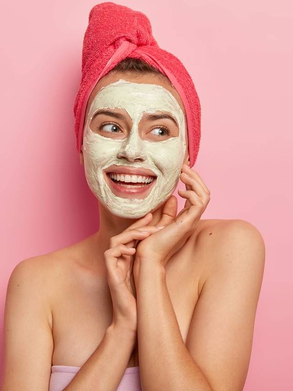 Isolated shot of pretty smiling woman with clay mask, makes beauty step, cleans face, wears wrapped towel on head, stands shirtless, gets pleasure, reduces pimples, copy space area against pink wall