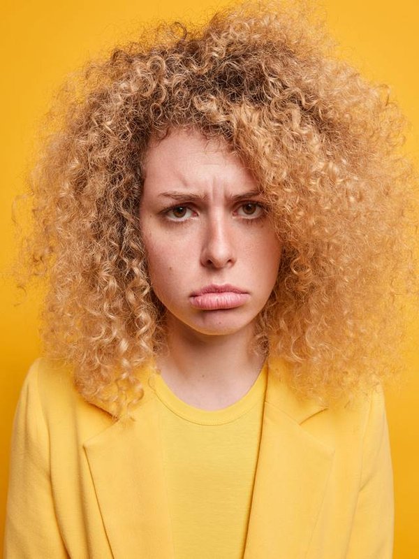 Portrait of sad unhappy young European woman with natural curly hair sulks from something unfair being offended purses lips expresses negative emotions has sorrowful disappointed expression.