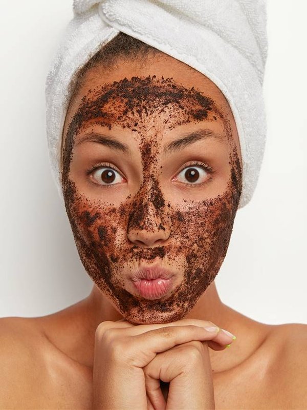 Portrait of attractive woman with surpised expression, has coffee scrub around face, cleanses from pores, removes dead cells, chooses mask to suit her skin, stands topless after shower, towel on head
