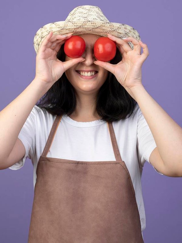 smiling young brunette female gardener in uniform wearing gardening hat covers eyes with tomatoes isolated on purple background with copy space