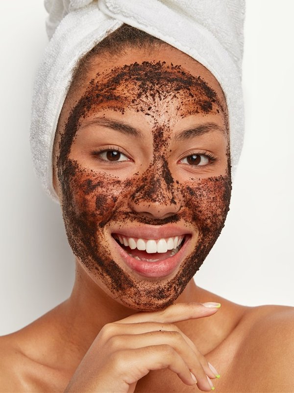 Close up portrait of happy Afro woman touches chin gently, smiles broadly, shows white teeth, cleans face, applies coffee scrub mask, wears wrapped towel on wet hair after taking bath. Skin care