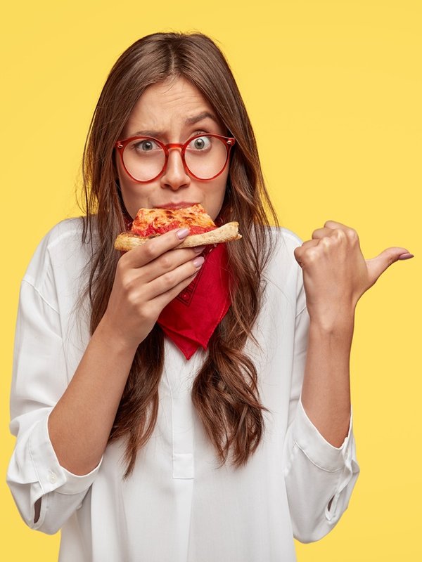 Horizontal shot of surprised female eats tasty slice of pizza, dressed in fashionable clothes, indicates with thumb, invites you to pizzeria, isolated over yellow studio wall. People and nutrition