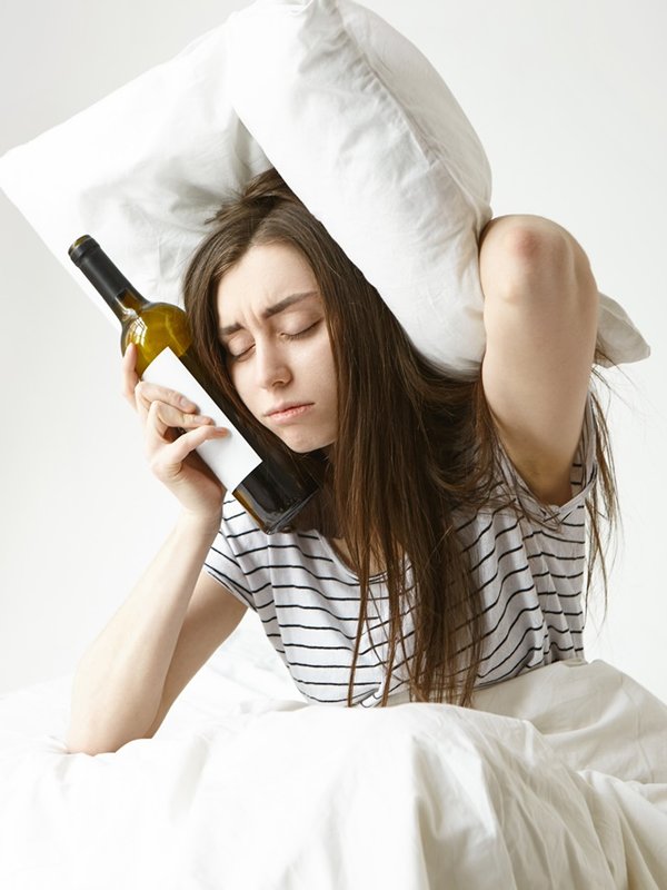 Sad young woman sitting on bed suffering from bad hangover after night party at club, having sleepy tired look, keeping eyes closed, holding bottle of wine and pillow, trying to cover ears from noise
