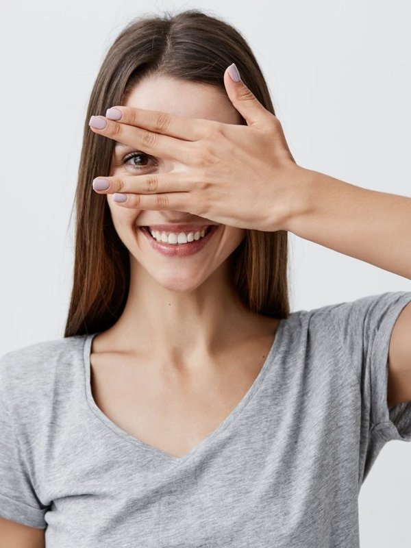 Close up portrait of beautiful joyful young caucasian student girl with dark long hair in trendy gray t shirt smiling with teeth, clothing eyes with hand, looking through finger with one eye.
