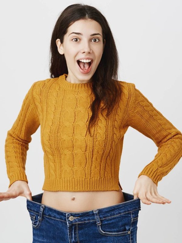 Astonished and happy young lady being impressed and excited because of losing weight, showing empty space in jeans by stretching it, standing over gray background. Girl is ready for summer