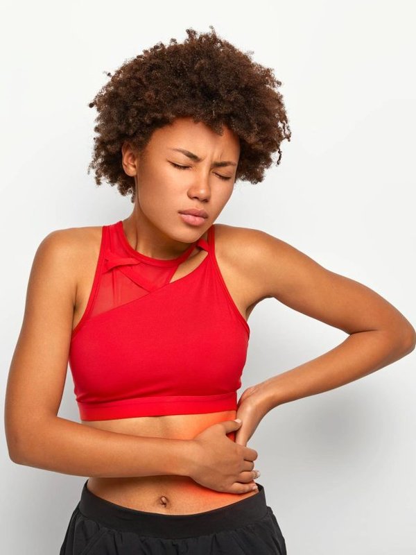 Dissatisfied African American woman holds aching hip, has kidney inflammation, touches location of pain near ribs marked with red dot, wears sport bra, shows perfect figure, has sporty shaped body