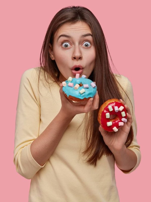 Astonished young cute European woman birtes sparkled delicious doughnut, keeps eyes wide opened, dressed in yellow sweater, has tasty snack, isolated over pink background. Sweet dish, nutrition