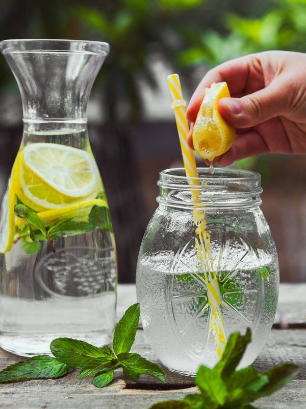 Hand squeezing lemon into a glass jar with water side view on wooden and yard background