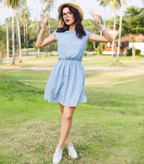 Young girl wearing sky-blue dress is standing on tip-toes in a park. Girl has straw hat and black glasses on