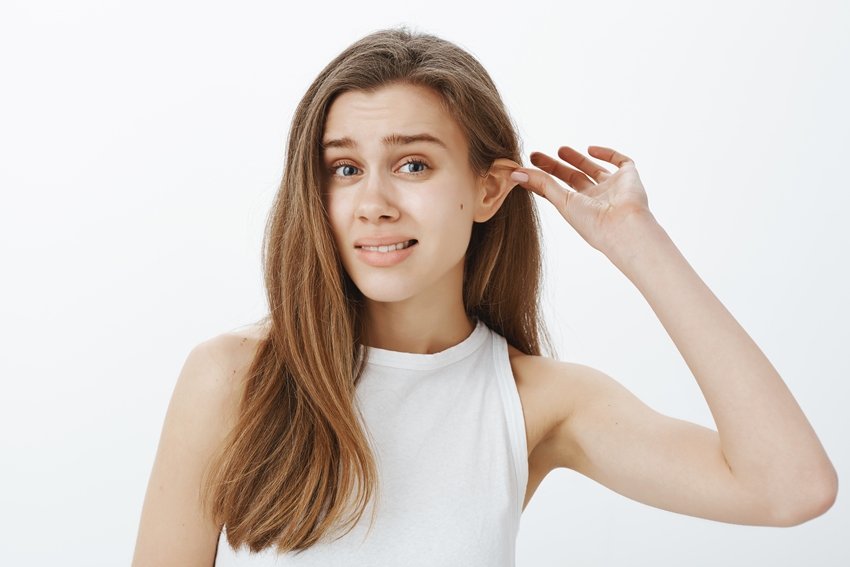 Stop lying my ears tired. Portrait of funny attractive woman with fair hair, pulling ear with hand and staring displeased at camera, feeling awkward while standing against gray background