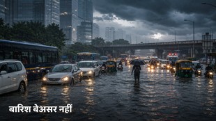 दिल्ली की एक प्रमुख सड़क पर भारी बारिश के बाद घुटनों तक जलभराव है। सड़क पर कारें, बसें, ऑटो-रिक्शा और मोटरसाइकिलें ट्रैफिक में फंसी हैं, जबकि एक व्यक्ति छाता लेकर पानी के बीच से गुजर रहा है। पृष्ठभूमि में आधुनिक इमारतें, ओवरपास, घने काले बादल और बारिश के बीच चमकती इमरजेंसी लाइटें दिखाई दे रही हैं।