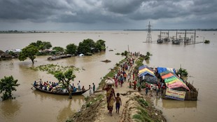 बिहार के बाढ़ग्रस्त इलाके में पानी से घिरे गांव, नावों और ऊंचे तटबंध पर सुरक्षित स्थान की ओर बढ़ते लोग।